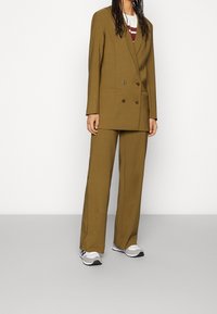 Brun dubbelknäppt kavaj med matchande vida byxor. Materialet verkar vara strukturerat; har två knappar och minimalt med detaljer. Bärs med grå sneakers.