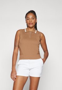 Brun ribbet uten ermer topp med hvit krage og frontlås, kombinert med hvite shorts. Strukturert stoff og tettsittende design.