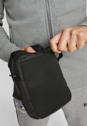 Musta verkkomäinen kassi vetoketjulla, suorakulmainen muoto, verkkohihna. Rakenteellinen pinta, jossa näkyvät saumat ja korostukset.