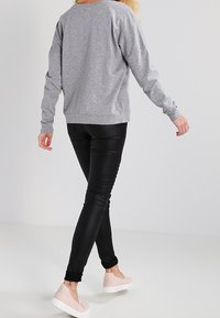 Persona che si allontana indossando una maglietta grigia a maniche lunghe, pantaloni neri aderenti e scarpe slip-on rosa chiaro su uno sfondo neutro.