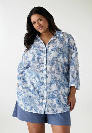 Donna con camicia azzurra a fantasia paisley, abbottonata, con maniche a 3/4, e pantaloncini blu su sfondo neutro, con le mani nelle tasche.