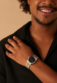 Armbanduhr in Gold- und Silbertönen mit blauem Zifferblatt, metallischem Armband und strukturiertem schwarzen Hemd. Auffällige Details sind eine geriffelte Lünette und Indexmarkierungen.