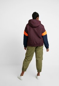 Chaqueta con capucha en color marrón y azul marino, con una franja naranja, que cuenta con puños elásticos y un corte holgado, combinada con pantalones cargo en color oliva y zapatillas blancas.