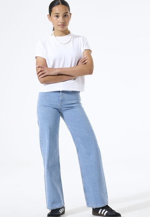 Camiseta blanca de manga corta combinada con vaqueros acampanados de tiro alto en azul claro y zapatos negros con rayas blancas. Diseño de conjunto sencillo y casual.