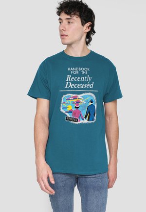 Young man wearing a teal t-shirt with "Handbook for the Recently Deceased" text and Beetlejuice-themed graphic of two figures holding hands.
