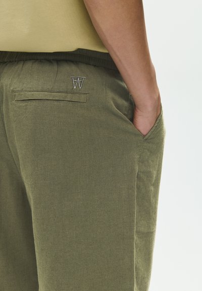 Personne portant un pantalon vert olive avec une ceinture élastique et la main dans la poche latérale, haut beige visible au-dessus du pantalon.