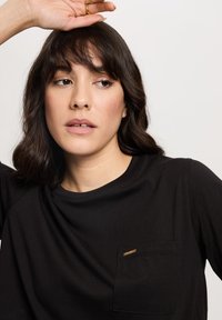 Czarna koszulka typu t-shirt z krótkim rękawem, wykonana z miękkiego materiału, z kieszonką na piersi ozdobioną małym złotym akcentem. Prosty, okrągły dekolt.