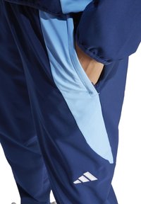 Marineblauwe sportieve joggingbroeken met lichtblauwe zijpanelen, een zak op de heup en een logo met drie witte strepen aan de onderkant. Gladde stofstructuur.
