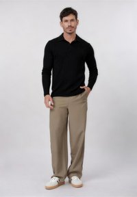 Άνδρας που φοράει μαύρο πόλο με μακριά μανίκια, μπεζ παντελόνι φαρδιάς γραμμής και λευκά αθλητικά παπούτσια, στέκεται με το ένα χέρι στην τσέπη, σε απλό φόντο.