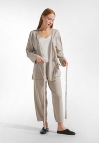 Set da salotto beige con un cardigan lungo e aperto con tasche, pantaloni larghi e una canottiera bianca, completato da scarpe slip-on nere.