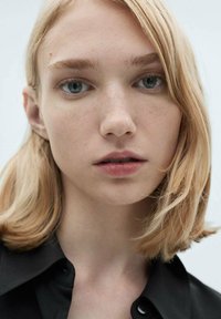 Portret osoby z jasnymi blond włosami i niebieskimi oczami, ubranego w czarną koszulę na guziki, na tle w neutralnych kolorach.