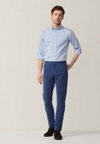 Lichtblauw button-down overhemd met opgerolde mouwen, gecombineerd met slim-fit donkerblauwe broek en donkerbruine schoenen op een neutrale achtergrond.