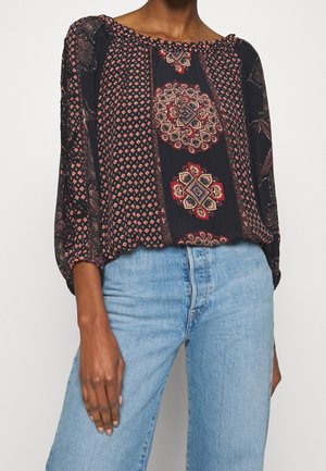 Frau trägt eine lockere schwarze Bluse mit roten und beigefarbenen geometrischen Mustern und hellblaue, hoch taillierte Jeans, steht vor einem weißen Hintergrund.