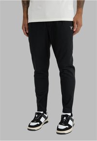 Musta urheiluhousut kapealla leikkauksella, joissa vasemmalla puolella kirjailtu logo, yhdistetty mustavalkoisiin tennareihin.