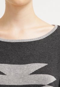 Gros plan sur une personne portant un pull gris foncé avec un motif géométrique gris clair et un large décolleté.