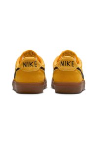 Sneakers in suede giallo con accenti neri, caratterizzati da una punta arrotondata e una suola in gomma naturale. Il tallone presenta in modo prominente il logo "NIKE".