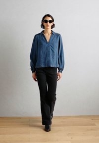 Løs blå denimbluse med v-hals, rynket frontsaum og lange ermer, kombinert med svarte rette bukser og spisse støvler.