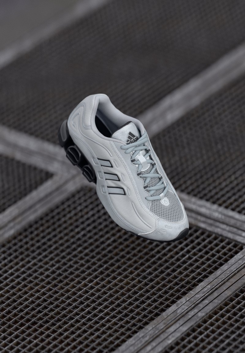 Light gray athletic shoe with mesh upper, three black stripes, rounded toe, and cushioned black sole. Features lacing and logo on tongue.