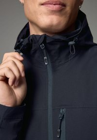 Giacca isolante scura con colletto alto, cappuccio regolabile e tasche con zip. Presenta una superficie testurizzata e una cerniera a contrasto.