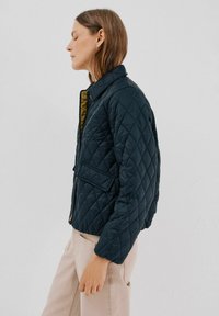 Marineblå quiltet jakke med krave, lynlåslukning og forlommer, med en kontrasterende gul indvendig side og struktureret stof.