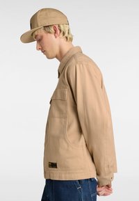 Beige canvas jacket with a collar, single chest pocket, and two side pockets; paired with a matching tan cap. Model facing left.