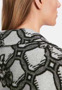 Grauer Stoff mit einem schwarzen geometrischen Muster, das florale Akzente aufweist. Weiche Textur mit einem geschmeidigen Fall, der auf die Details des Musters und die Form fokussiert.