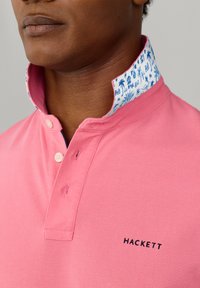 Gros plan sur une personne portant une chemise rose à col avec des boutons et un col intérieur blanc décoré d'imprimés tropicaux bleus.