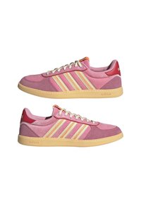 Paire de baskets Adidas roses avec des bandes crème, des semelles beiges, des lacets blancs et des accents rouges au talon, vues de côté sur fond blanc.