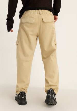 Person trägt beige Cargohose mit Klappe am Rücken und Seitentaschen, schwarzen Langarmpullover und schwarze Sneakers mit weißen Details, von hinten betrachtet.