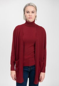 Rød cardigan i blød strik, åben front design med sidelommer. Laget over en matchende turtleneck, suppleret af blå jeans.