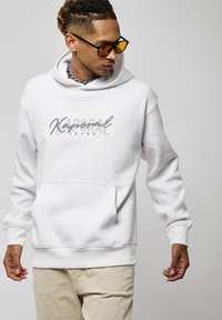 Hoodie blanc avec une poche frontale et un logo "Kaporal Jeans" en bleu et beige. Le tissu semble doux avec une texture lisse.