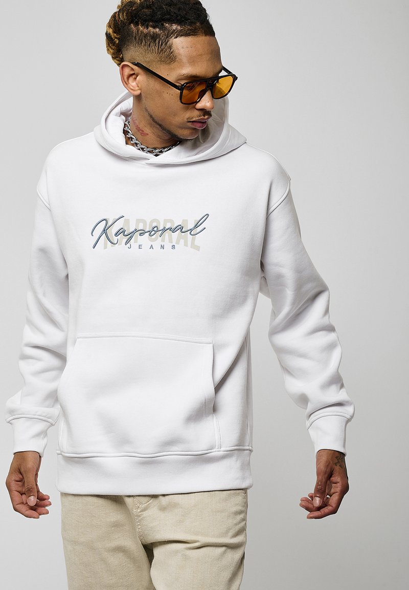 Hoodie blanc avec une poche frontale et un logo "Kaporal Jeans" en bleu et beige. Le tissu semble doux avec une texture lisse.