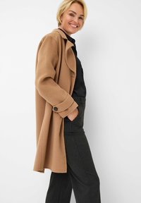 Beige Wollmischungsmantel mit lockerer Passform, übergroßen Revers und schwarzen Knopf-Accents, getragen über einem schwarzen Hemd mit Knöpfen und dunkel gestreiften Hosen.