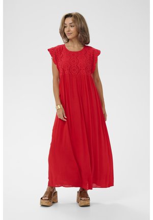 Kobieta w długiej czerwonej sukience z koronkowymi detalami na górze, stojąca z luźno opuszczonymi rękami, w brązowych sandałach na platformie i złotej bransoletce.