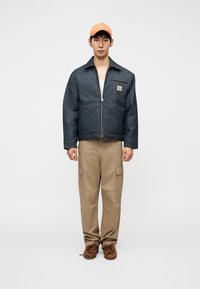 Τζιν μπουφάν με φερμουάρ, χακί παντελόνι cargo και καφέ παπούτσια· πορτοκαλί καπελάκι προσθέτει χρώμα στην εμφάνιση. Απλός σχεδιασμός, κλασικά χρώματα.