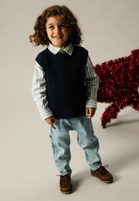 Niño con un chaleco de punto azul marino encima de una camisa de rayas clara, combinado con unos vaqueros azul claro y zapatos mocasines marrones. Decoración de papel de aluminio rojo cerca.