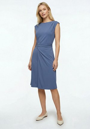 Femme portant une robe bleue sans manches, longueur genou, avec un détail de boucle latérale, debout devant un fond uni clair.