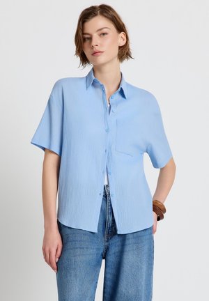 Giovane donna che indossa una camicia azzurra a maniche corte, ampia e sbottonata, e jeans blu a vita alta, in piedi con una mano sui fianchi.