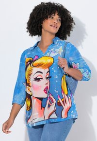 Femme aux cheveux bouclés portant une chemise bleue ornée d'une grande femme de dessin animé colorée en train de s'appliquer du rouge à lèvres, tenant un sac à main bleu brillant.