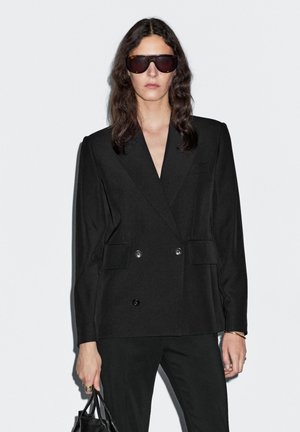 Blazer nero doppiopetto con colletto a revers, due tasche frontali e texture liscia. Indossato con pantaloni neri e con una borsa.