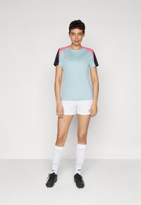 Camisa de fútbol azul claro con mangas negras y detalles en rosa, combinada con pantalones cortos blancos, botas negras y calcetas blancas hasta la rodilla con un patrón.