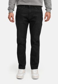 Mann iført svarte jeans med rette ben, grå genser og hvite joggesko, stående foran en hvit bakgrunn.