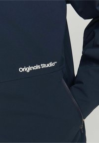 Giacca blu navy con una texture liscia, dotata di una tasca con cerniera e il logo "Originals Studio™" bianco sul davanti.