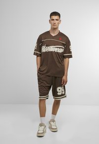 Barna sportos öltözet, amely v-nyakú mezből áll, fehér felirattal és rövidnadrágból, amelyen nagy '99' szám szerepel. Puha anyag, csíkos díszítéssel.