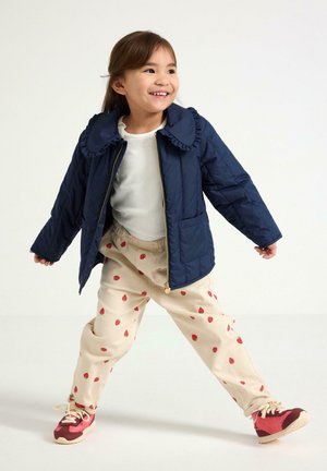 Laineissa tikkikangas takki, jossa on röyhelökaulus, kermansävyiset housut, joissa on hajallaan punaisia mansikkakuviota, sekä punaiset ja vaaleanpunaiset lenkkarit.