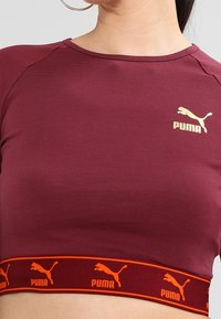Burgunderfarbenes, tailliertes Crop-Top aus glattem Stoff mit einem goldenen Puma-Logo auf der Brust und einem orangen Logo-Band am Saum.