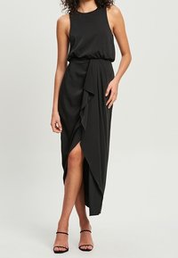 Vestido negro sin mangas con detalle de falda drapeada y abertura lateral, confeccionado en tejido suave, que presenta un ajuste relajado y una textura suave.