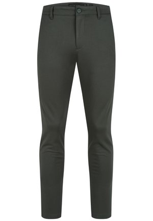 Mężczyzna ubrany w czarne wąskie spodnie, białą koszulkę z okrągłym dekoltem i białe trampki, stojący na tle białego, gładkiego tła.