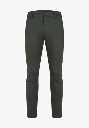 Homme portant un pantalon noir ajusté, un t-shirt blanc à col rond et des baskets blanches, debout devant un fond blanc uni.