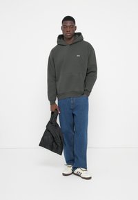 Grå hættejakke med frontlomme og små bogstaver, parret med blå jeans og hvide sneakers. Holder en sort jakke i den ene hånd.